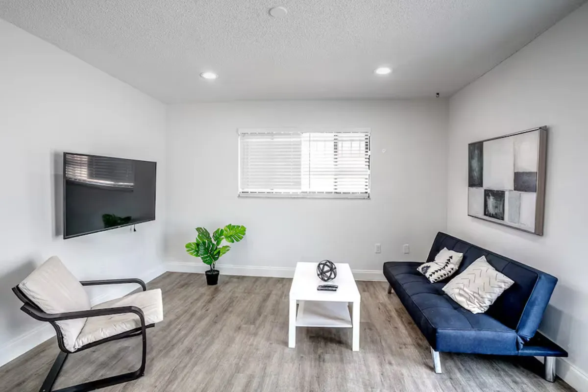 The hero image showcases a sunlit, contemporary living room featuring a plush blue modern sofa adorned with stylish patterned cushions, a sleek white coffee table with a decorative accent, a large wall-mounted Smart TV, and a vibrant indoor plant beside a window that lets in abundant natural light. The scene exudes comfort, modernity, and an inviting, tranquil atmosphere despite being in the heart of Miami.