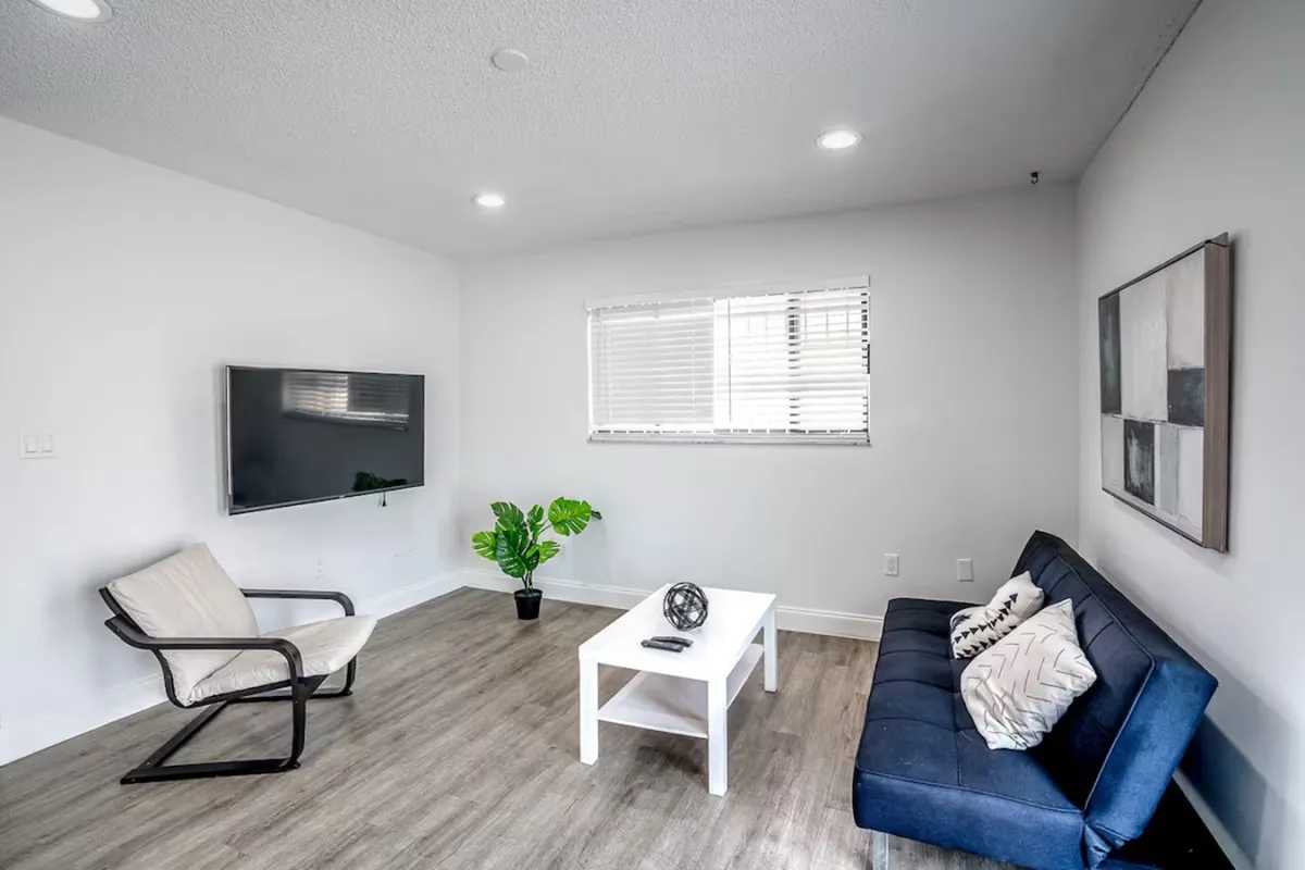 The image depicts a cozy living room featuring a wall-mounted Smart TV, a navy blue sofa with patterned pillows, a white coffee table with a decorative object, and a single armchair with a light cushion. The setting is relaxed and comfortable, offering a perfect spot for unwinding or enjoying entertainment after a day out in the city.