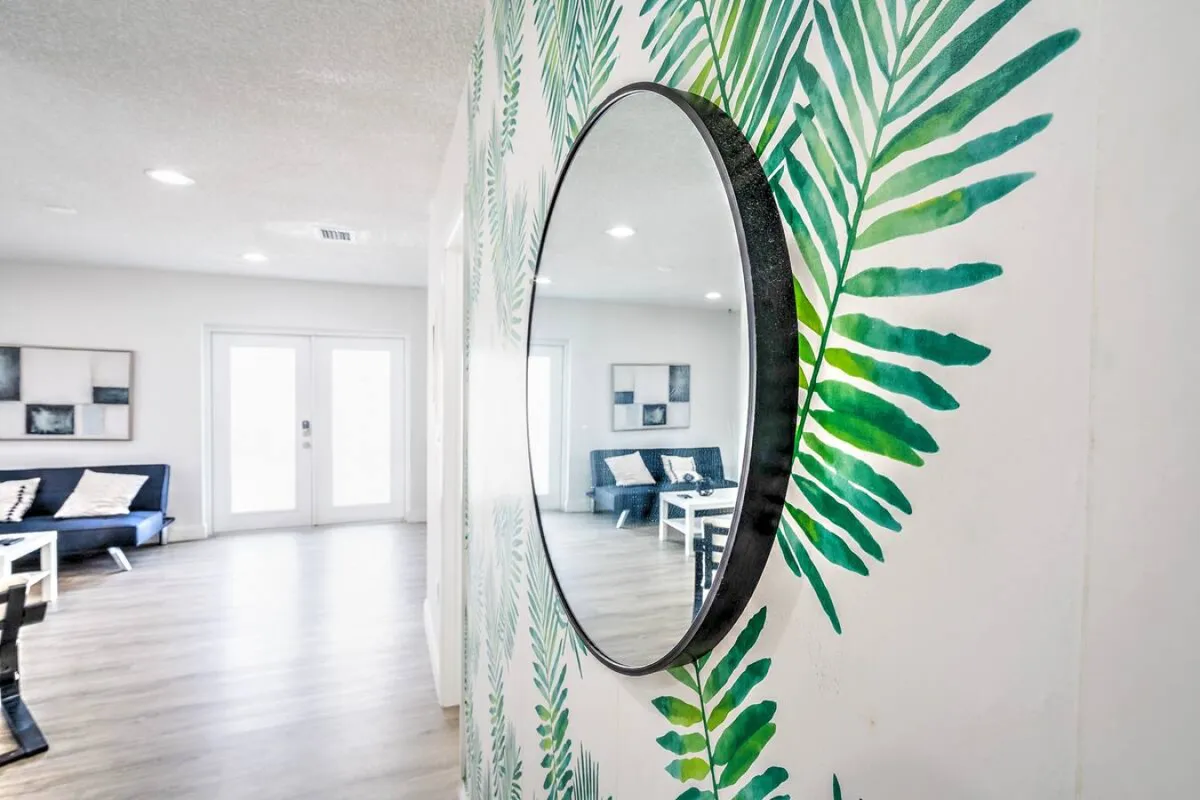 A bright, inviting living room with a large round mirror, tropical leaf patterned wallpaper, a comfortable blue sofa with cushions, and a white coffee table. Large doors likely lead to an outdoor space, allowing natural light to flood the room and creating a seamless connection between the indoors and the vibrant city outside.