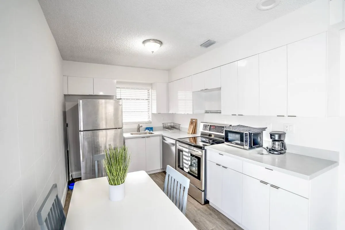 The kitchen is the focal point here, with a stainless steel refrigerator, stove with oven, microwave, and a small dining table with a plant centerpiece. The inviting and fresh ambiance makes this a practical space for preparing meals, enjoying morning coffee, or casual dining.
