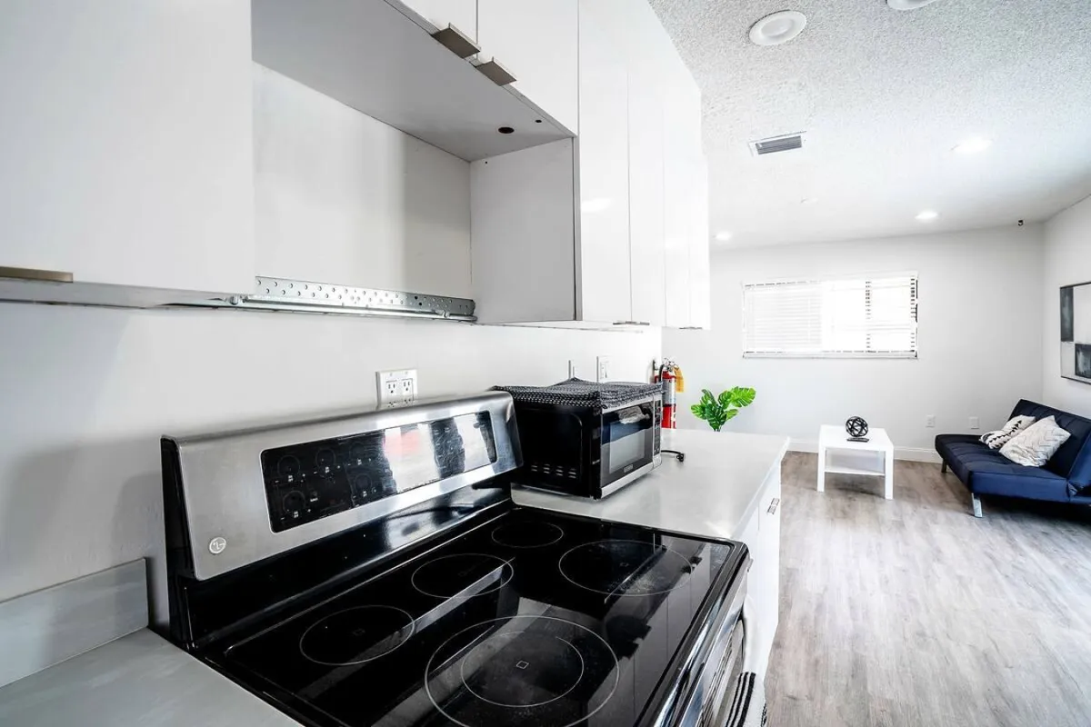 This perspective showcases the open-plan layout, with a view of the modern kitchen featuring an electric stove with glass cooktop, microwave on the countertop, minimalist white kitchen cabinets, and a glimpse of the living area with a dark blue sofa and white coffee table in the background. The space feels bright, airy, and contemporary.