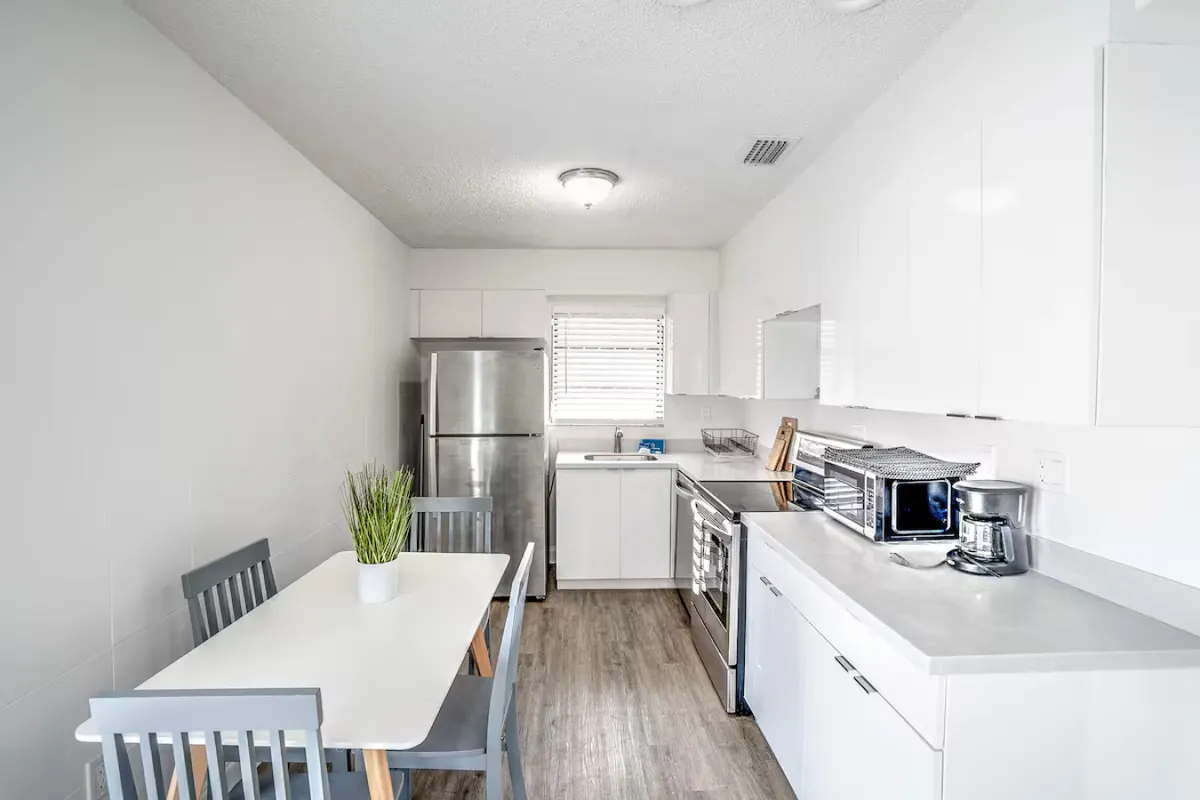 A modern kitchen and dining space with a white rectangular table, potted plant centerpiece, stainless steel refrigerator, electric stove and oven, and sleek white cabinetry. The scene is bright, tidy, and practical.