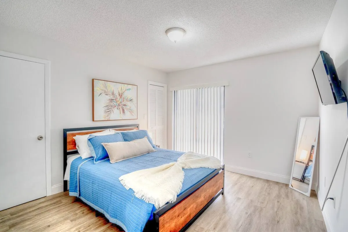A bright and airy bedroom featuring a neatly made bed with blue bedding and decorative pillows, a wall-mounted flat screen TV for entertainment or business presentations, a full-length mirror for getting ready, and tasteful framed artwork above the bed. Natural light suggests an inviting atmosphere ideal for both rest and informal work.