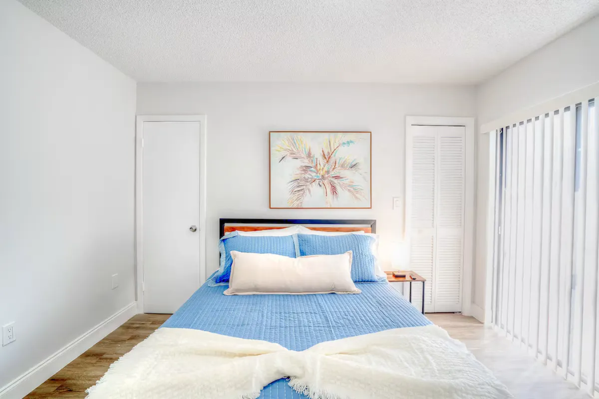 This interior shot looks out from a relaxing bedroom with a blue quilt and framed artwork, through sliding glass doors with vertical blinds that lead to the terrace. The scene hints at the transition from restful indoor space to the inviting outdoor terrace, filled with natural light.