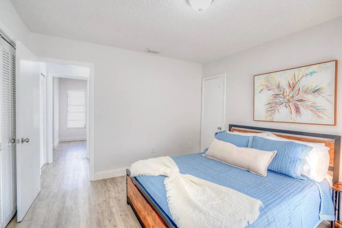 A peaceful bedroom featuring a king-sized bed with blue bedding and a beige throw pillow. Abstract palm tree artwork hangs above the bed, and an open door leads to a hallway filled with natural light. The room is finished with wood-style flooring, creating a clean, modern feel.