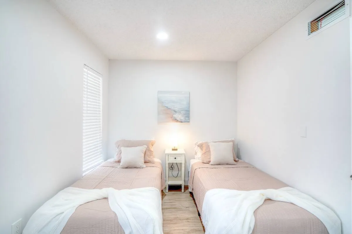 A cozy twin bedroom with two neatly made beds, beige bedding, a central white nightstand with a lamp, and soft beach-themed wall art. The space feels calm, organized, and inviting—ideal for guests seeking privacy and comfort.