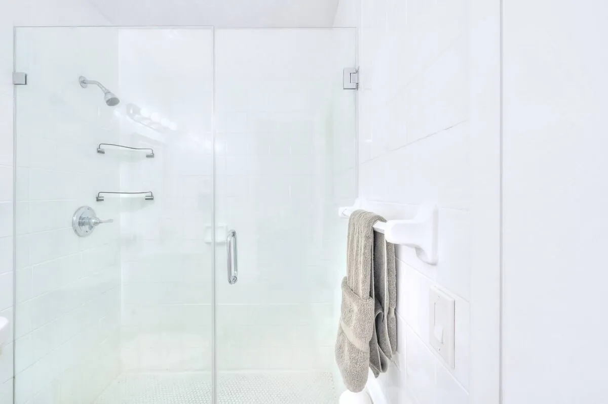 A bright, sparkling bathroom featuring a glass shower enclosure with chrome fixtures, a towel rack with beige towels, and crisp white tiled walls and floors. The space feels clean, modern, and inviting—offering everything needed for daily routines.