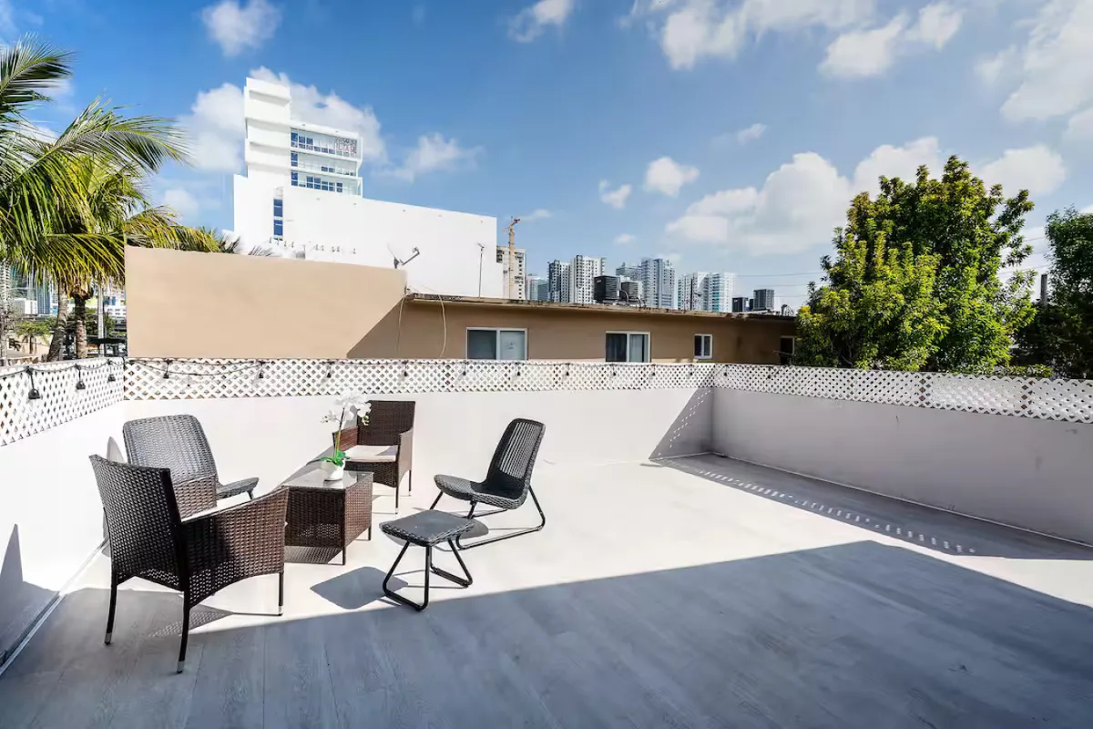 A cozy outdoor wicker seating set with a small table sits on a terrace framed by a white lattice fence. The distant Miami skyline and lush trees provide a vibrant urban backdrop, blending the tranquility of a private retreat with the energy of the city.