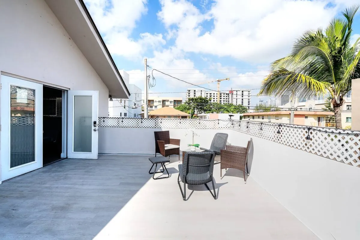 This image captures a private terrace with inviting outdoor seating, surrounded by a white lattice fence and accented by a palm tree. The urban skyline with mid-rise buildings is visible in the background, and the terrace is bathed in natural sunlight, creating a serene, open-air environment.