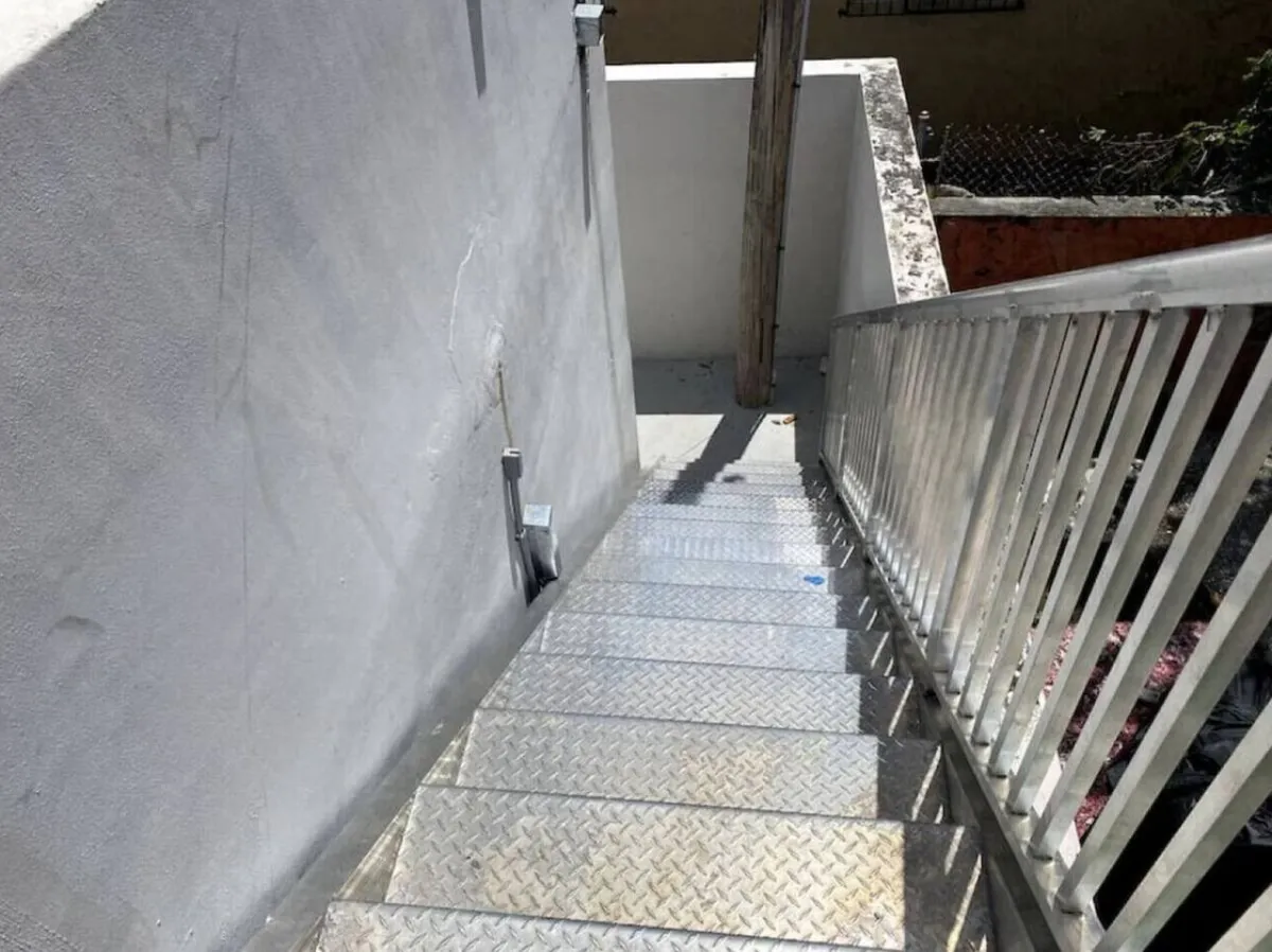 This image captures the building's exterior metal staircase with diamond plate steps, a sturdy white metal railing, and a concrete wall beside the stairs. The utilitarian design hints at the security and accessibility of the building's entry points.