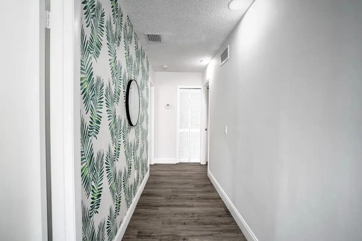 This interior hallway features tropical leaf patterned wallpaper, a round wall mirror, warm wooden flooring, and a white louvered door at the end. The scene feels fresh, calm, and welcoming, signaling the transition from the building entrance to the private spaces.